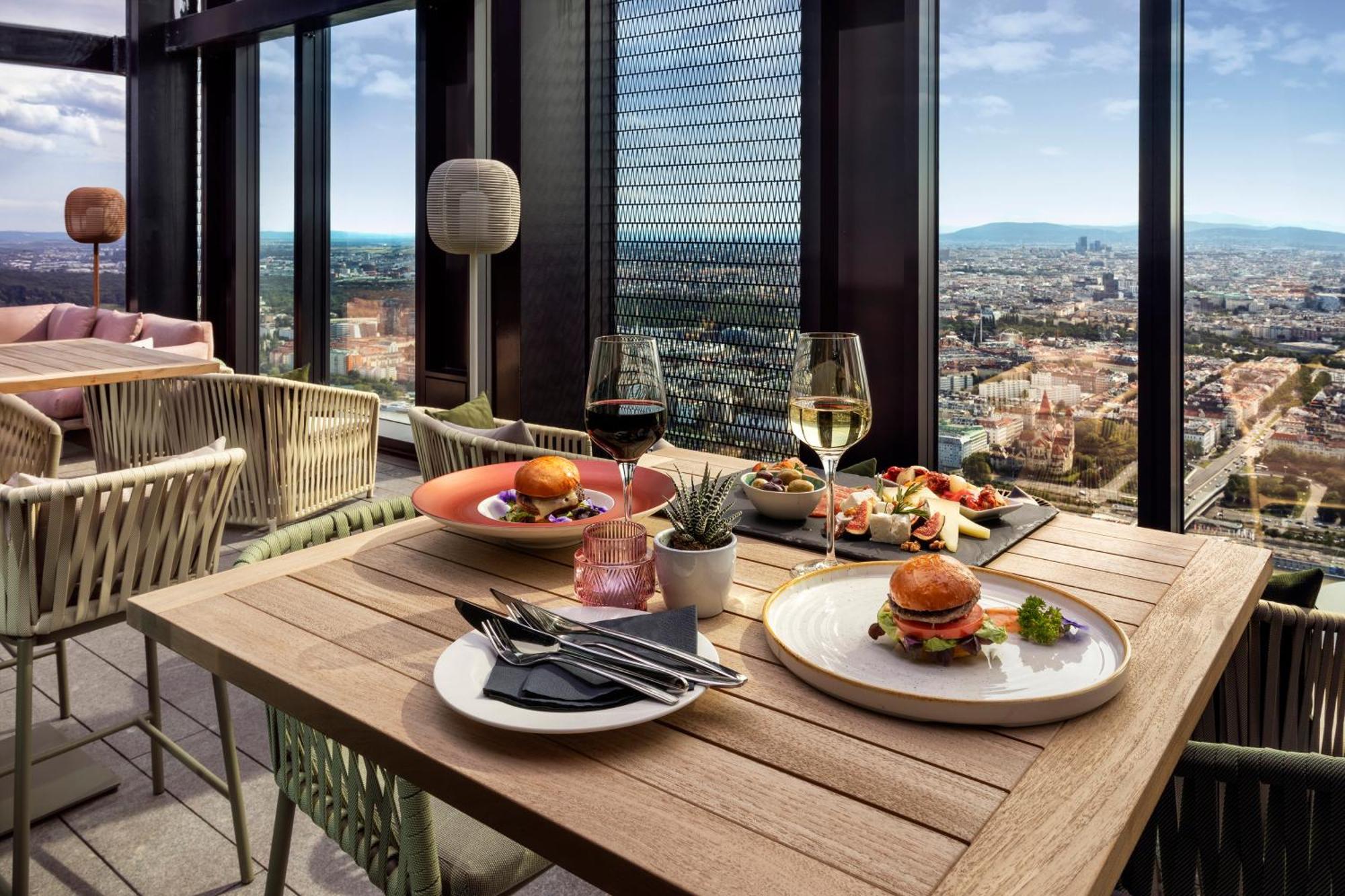 Melia Vienna Hotel Exterior foto The photo shows a stylish dining setup on a high rooftop or terrace, with a panoramic view of a city skyline in the background. 

There are two tables made of wood; one features two glasses of white wine and an assortment of dishes, including a plate