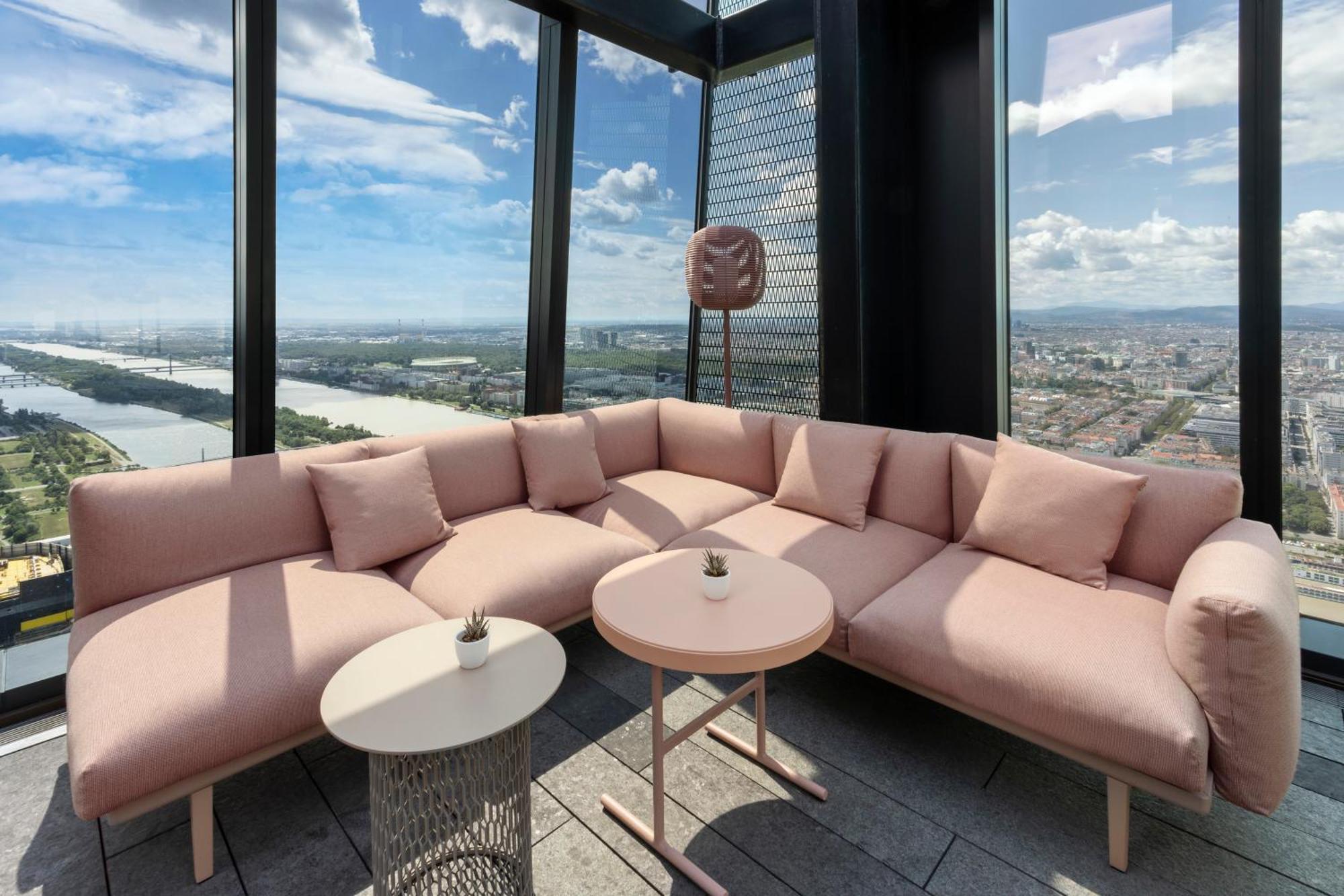 Melia Vienna Hotel Exterior foto The photo shows a modern lounge area with a large, L-shaped pink couch adorned with several soft cushions. In front of the couch, there are two tables: one round and one with a more intricate design. The space is located in a high-rise building, offe