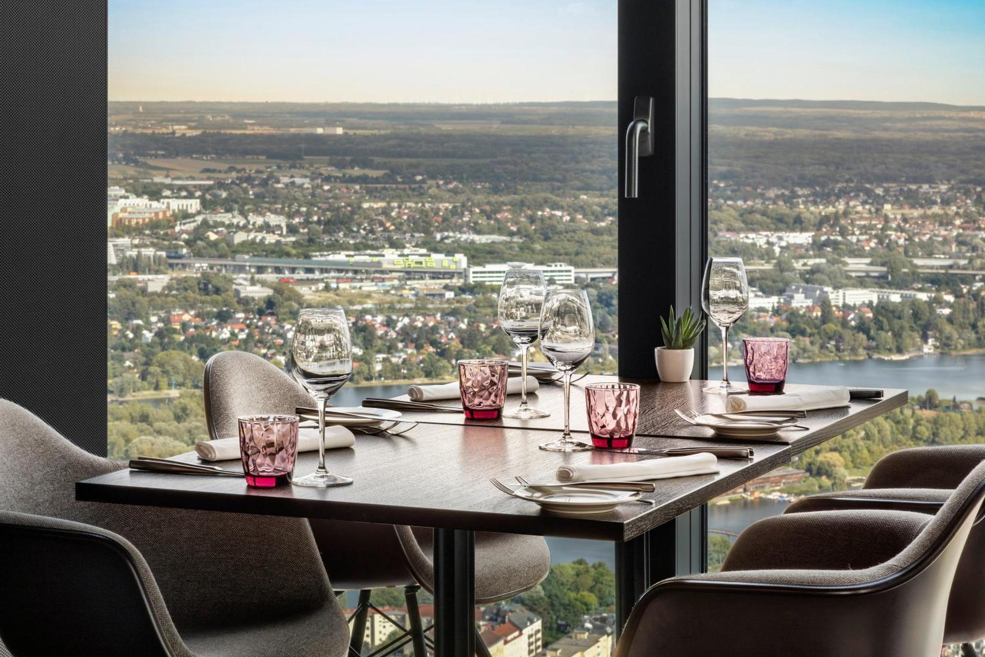 Melia Vienna Hotel Exterior foto The photo shows a dining table set up in a restaurant with a view. The table has dark wood and features several dining essentials arranged neatly. There are elegant wine glasses, pink-tinted glass holders, and plates set beside them. A small plant is